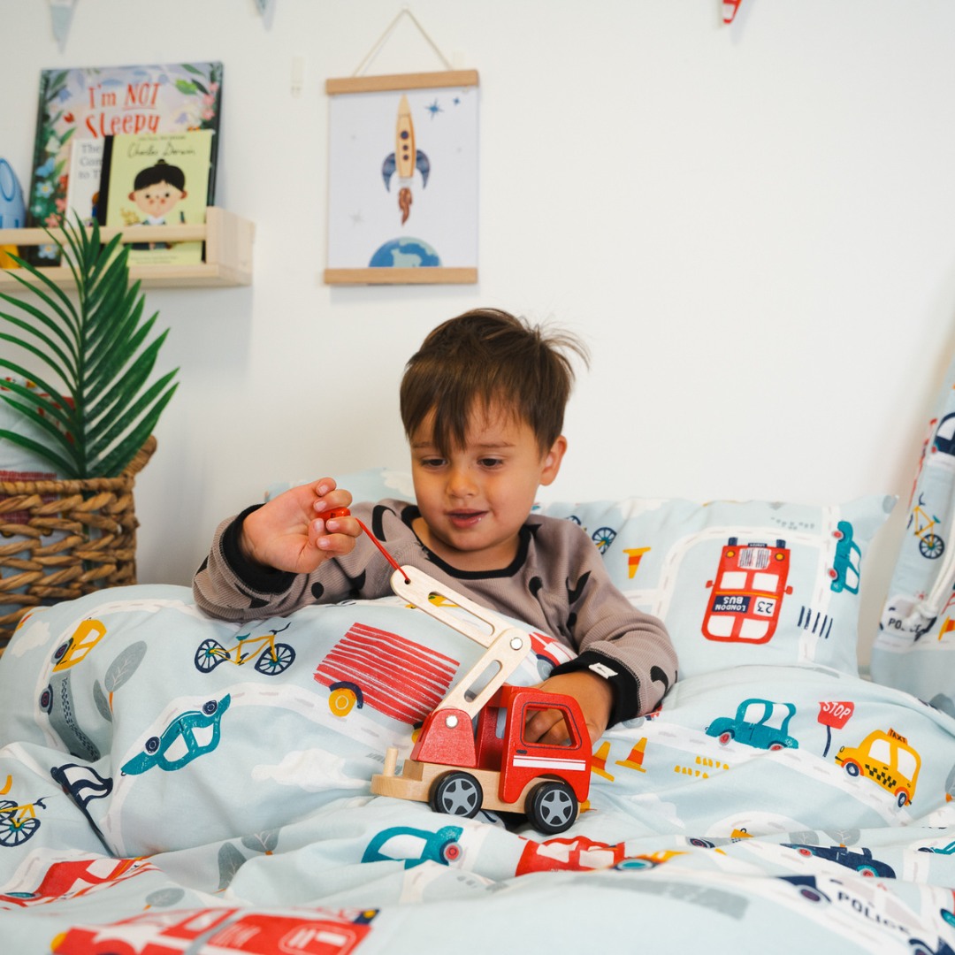 Young boy playing with a wooden toy truck in bed. The covers have colourful transport-theme illustrations: a bus, a taxi, cars, bikes. 