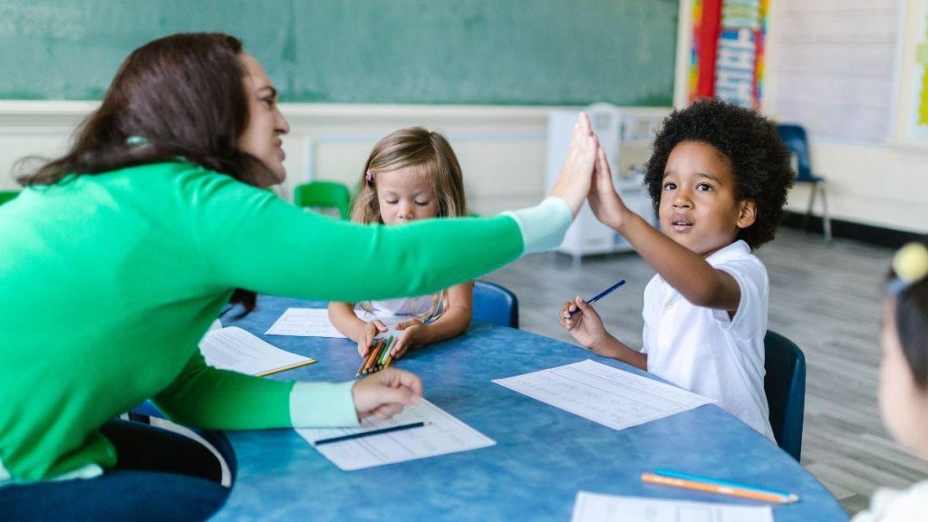 Teacher high-fives a student who is drawing a picture with another girl concentrating on her picture in the background.
