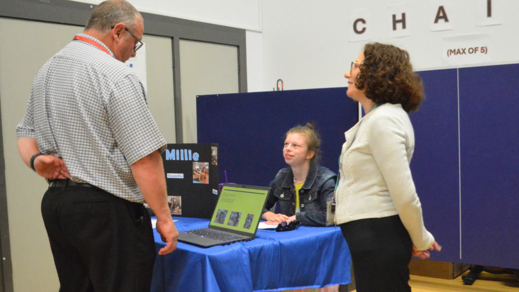 photograph of a student sat a table looking up at two people who are talking to her