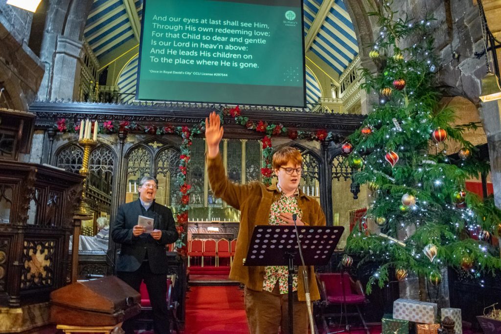 Teenage boy wearing a corduroy blazer singing. He is holding one hand up. He is stood in front of a music stand in an old church. Behind him is a screen with lyrics to the song.