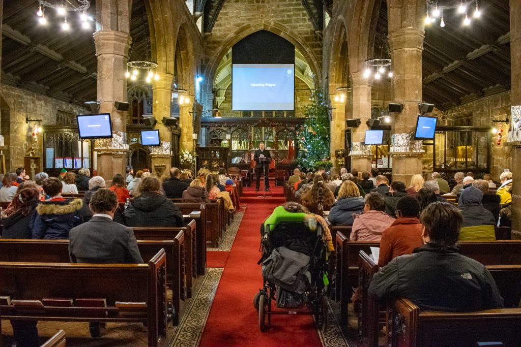 Old church illuminated and decorated for Christmas. There are people sitting down looking forwards at the pastor speaking.