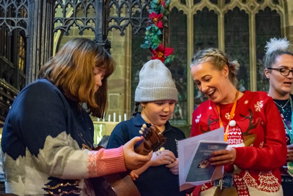 Two young girls and their teacher singing. One of the girls is holding a guitar and the other one is holding a sheet with lyrics.
