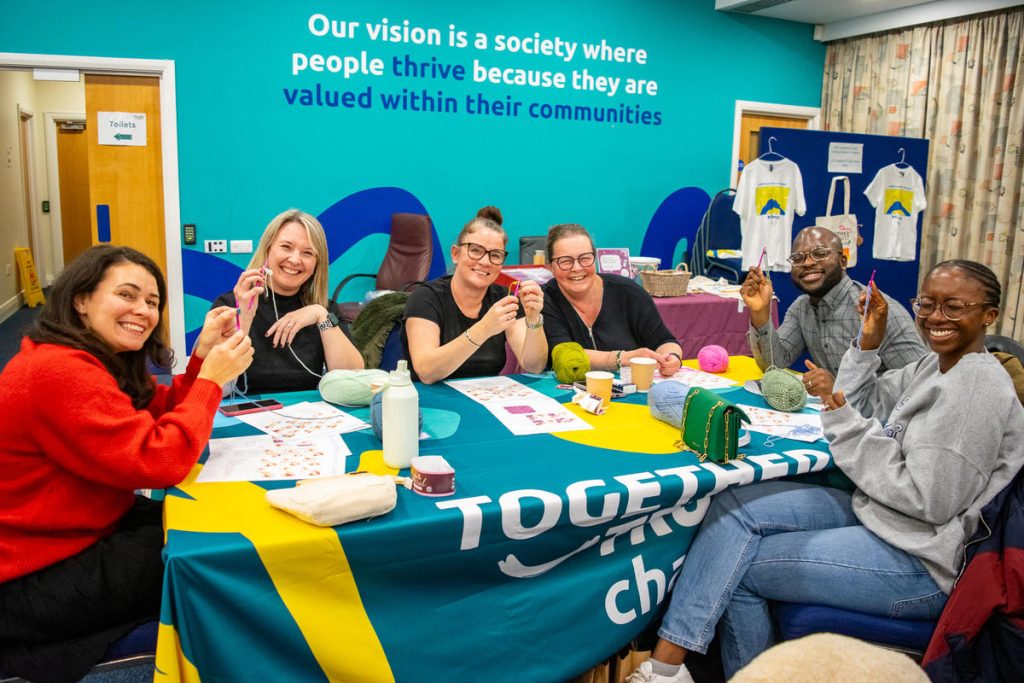 Six people sitting around a table smiling into the camera showing off their crochet creations.