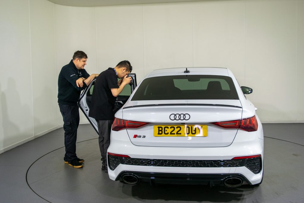 A student, teenage boy, is sitting in the front seat of a car smiling. He is holding and pointing a camera out. 
