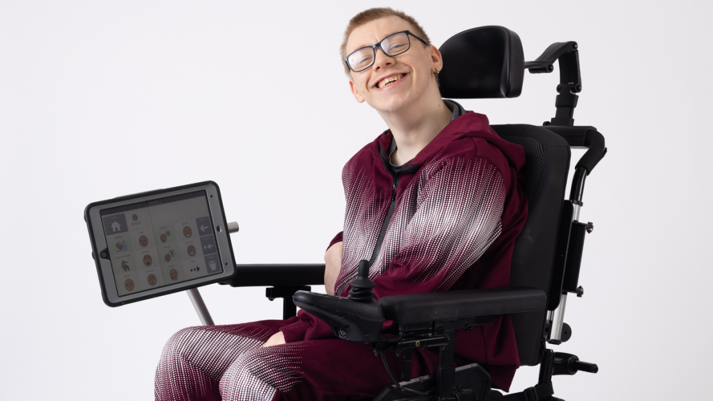 a young man sat in his wheelchair smiling at the camera. Attached to his wheelchair is an electronic communication device