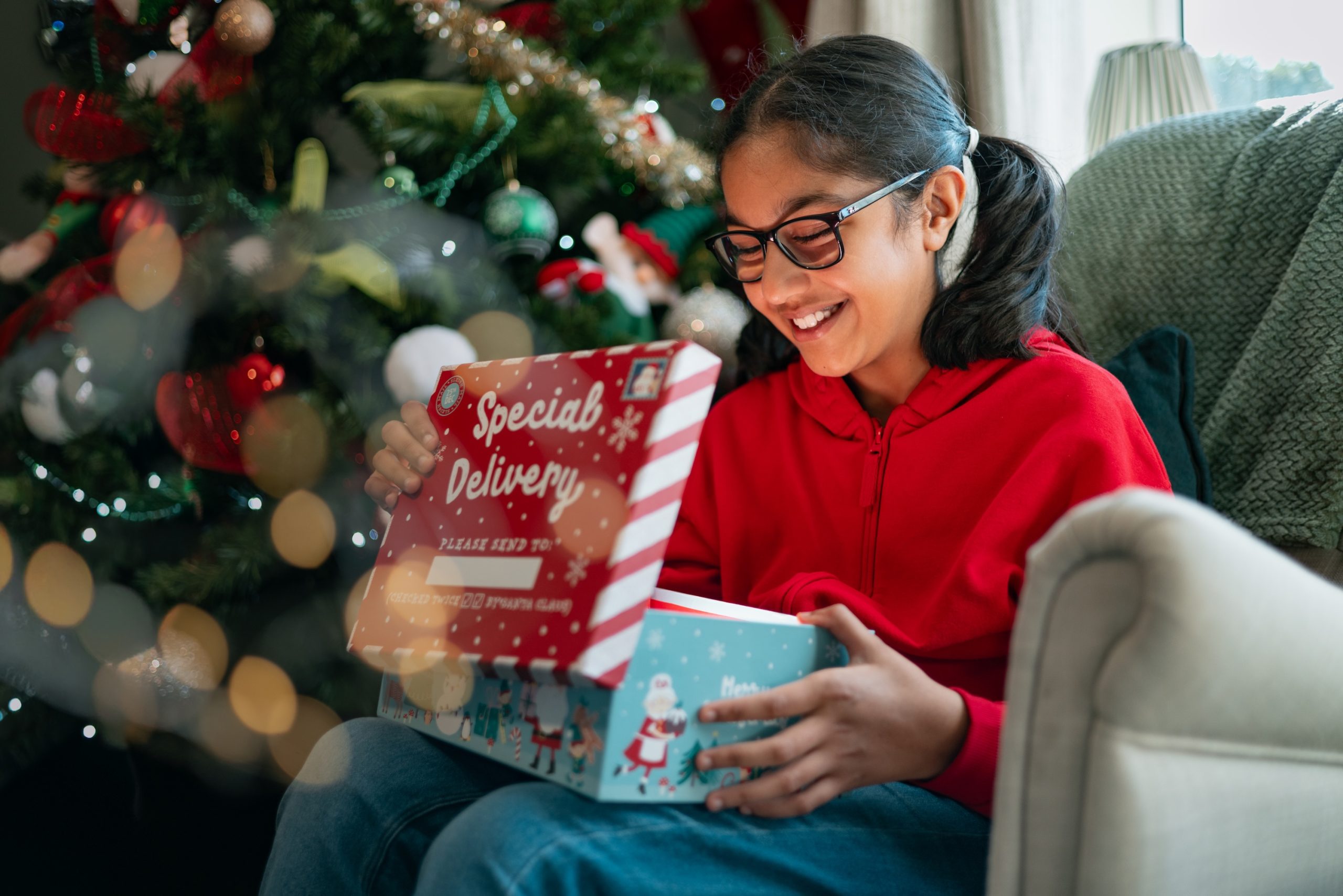 a girl in a red jumper sat in front of a Christmas tree opening a present
