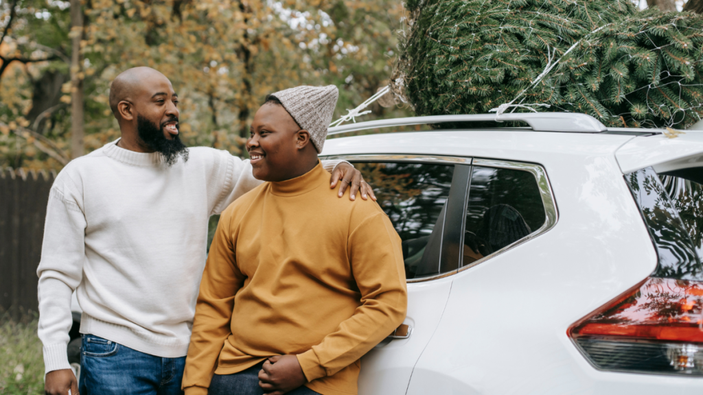 A man standing next to a teenager with one arm around his shoulder. The are outside leaning on a car. on top of the car is a christmas tree.