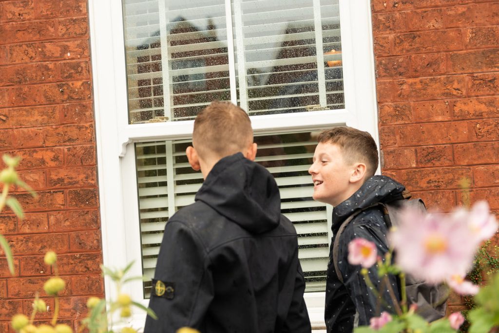 A photo of 2 boys walking and talking to each other past a window of a house