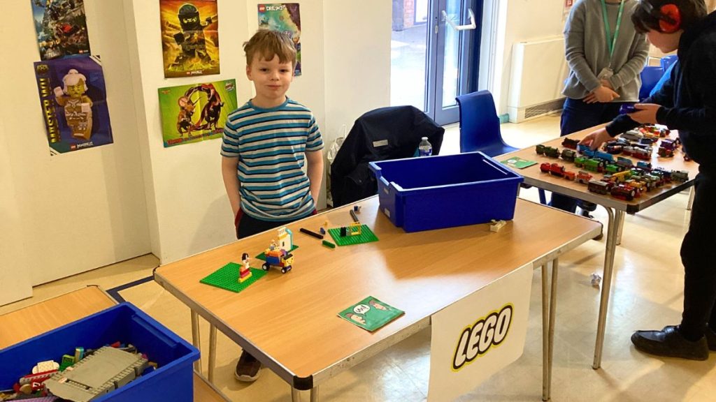 A child casually posing for a photo with hands tucked into their pockets, standing behind a stall adorned with the iconic 'Lego' sign.