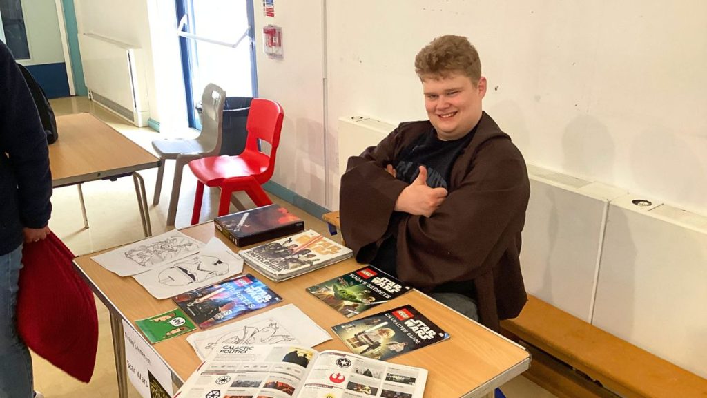 A genuine snapshot captures a cheerful young man, giving a thumbs-up to the camera from behind a stall adorned with a 'Star Wars' theme.