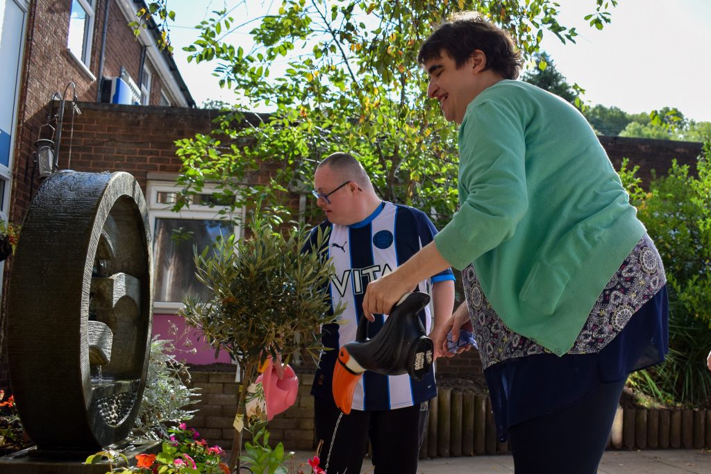Two adults, a woman and a man, in the day centre garden, both holding watering cans and watering some potted flowers.
