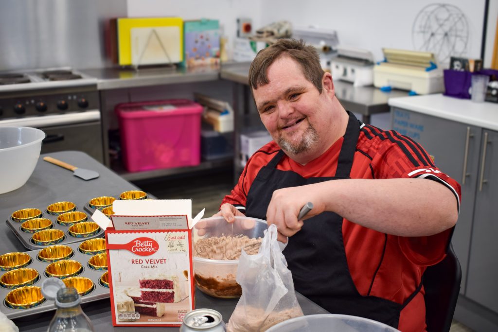 A man with down syndrome is sitting in an industrial style kitchen with an apron on and is stirring into a bowl of cake batter. He is smiling at the camera.