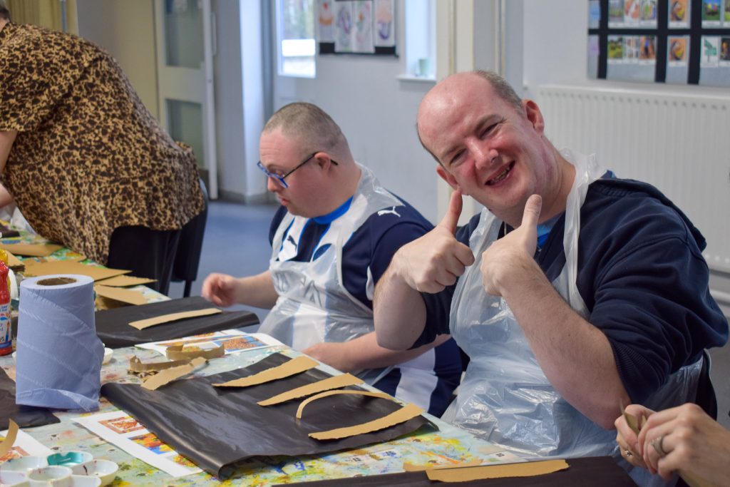 Two men are sitting down at a table wearing craft aprons. They are assembling a piece of art using craft materials.