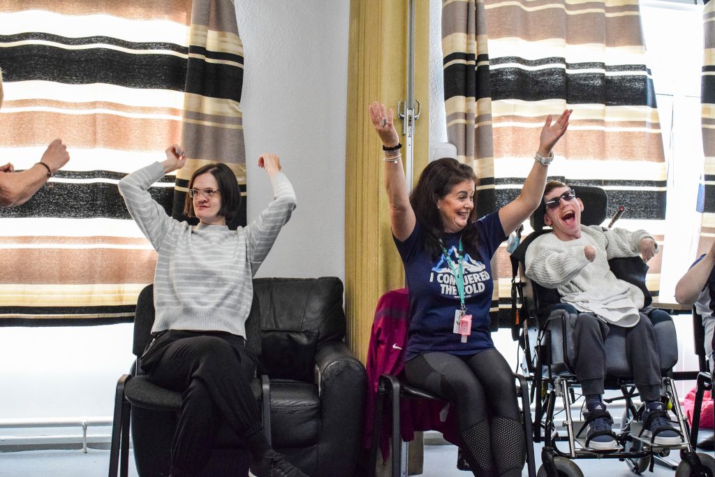 Three people, two women and a man, sit down on chairs dancing, two adults at the day centre and a staff member. One of the adults is in a wheelchair.