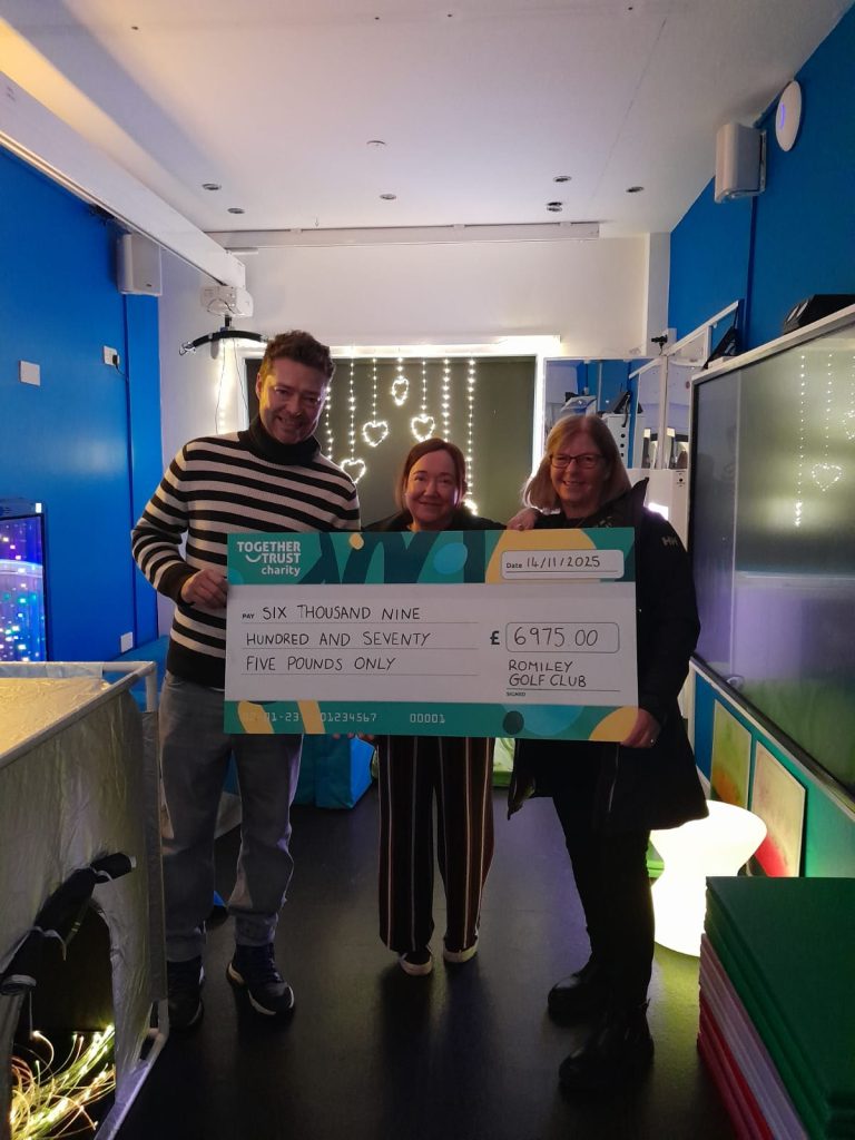 A group of three people holding a donation cheque that reads "£6975". two of them are from the romiley golf club, and one is a staff member at Woodlands.