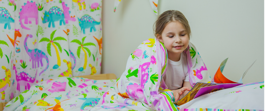 A girl is reading a book in bed. The betting matches the curtains behind her and depict a vibrant and colourful dinosaur print. 
