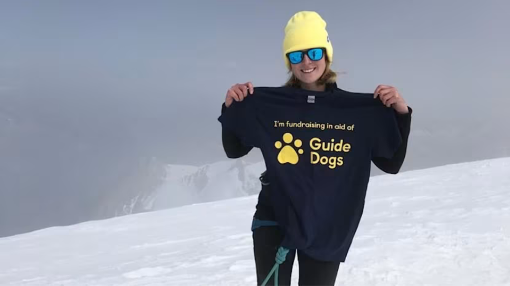 Lucy, a young woman wearing a yellow beanie, sunglasses and hiking clothes is holding up a T-shirt that reads "I'm fundraising in aid of Guide Dogs". She is posing on top of a mountain, with snow and distant mountains in the background behind her.