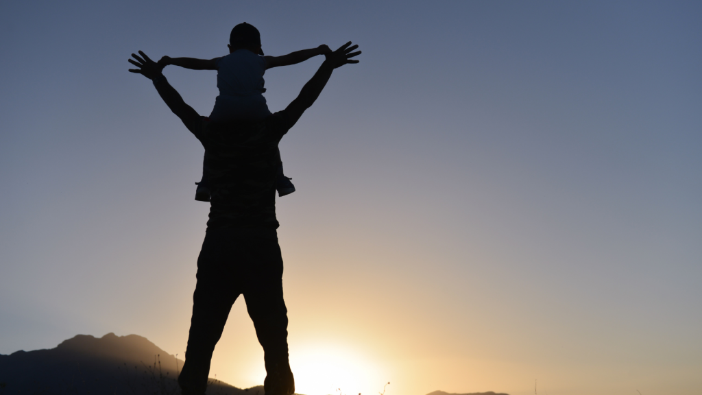 A silhouette of a man holding up a child on his shoulders against a blue sky.