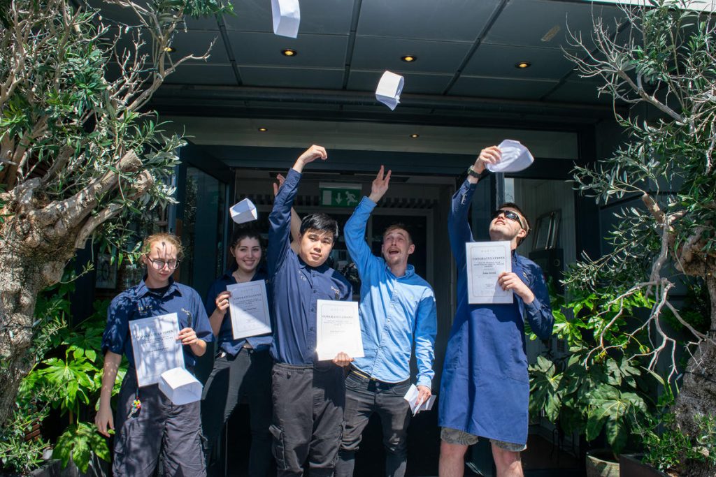 A group of 4 students and an employee from Gusto throwing up paper hats in the air outside the restaurant. 