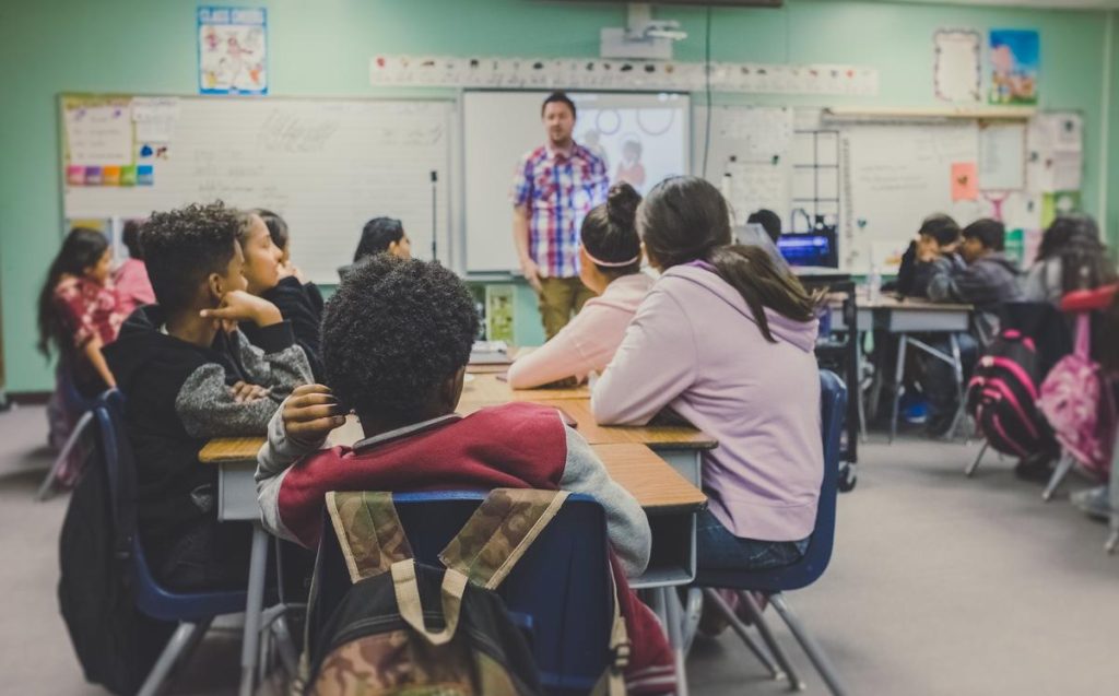 Photo of students in a class room with a teacher at the front speaking Image by kenny-eliason-zFSo6bnZJTw-unsplash