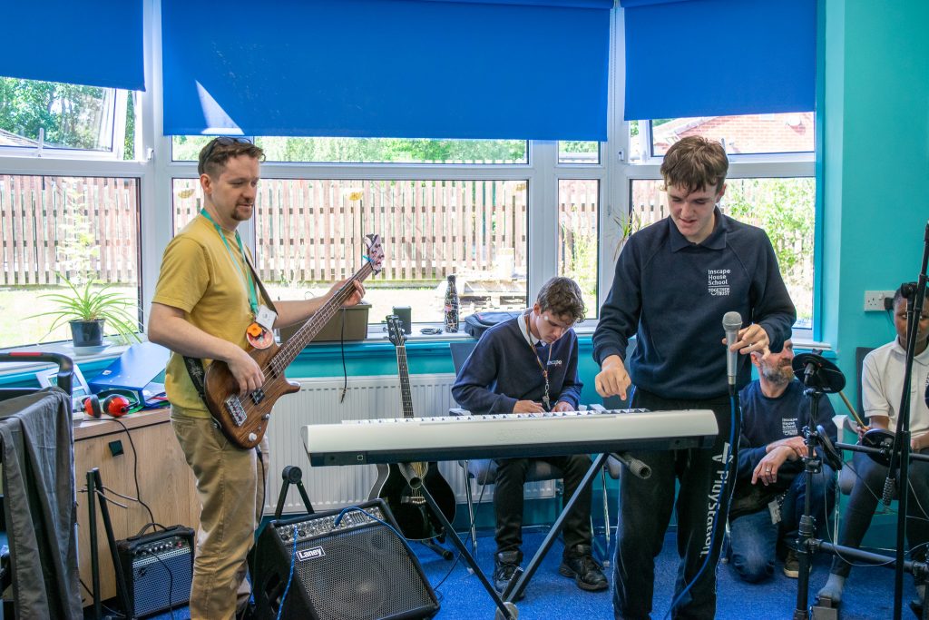 Music teacher holding a guitar playing music with 3 students: one on piano, one on drums and another singing. 