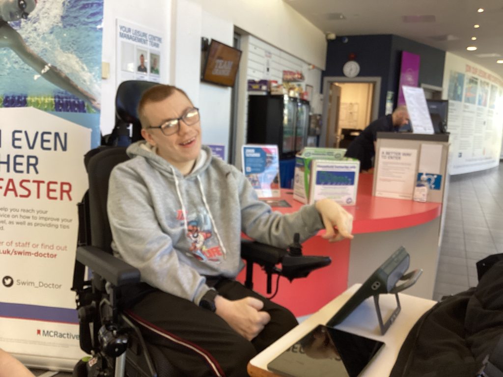Rio, a young man in a motorised wheelchair, smiles at the reception desk at Manchester Leisure Centre. 