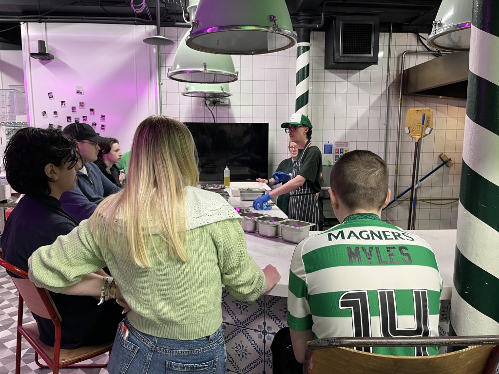 A group of students sitting around a circular countertop watching a pizza making demonstration. 