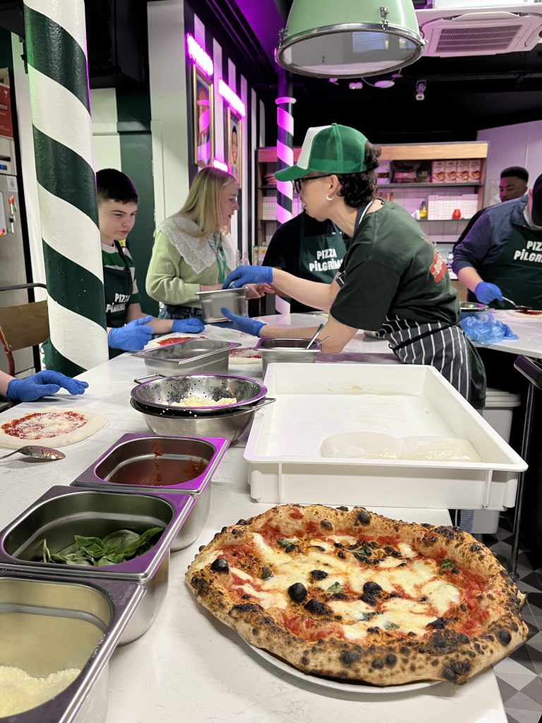 A pizza chef is leaning over a countertop to show students pizza toppings option. In the foreground, there is a freshly baked pizza. 