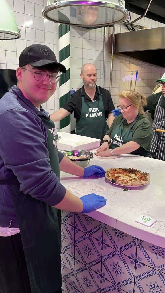 A young man proudly showcases the pizza he's just made. Behind him, 2 staff members from Pizza pilgrims look at the progress students are making. 