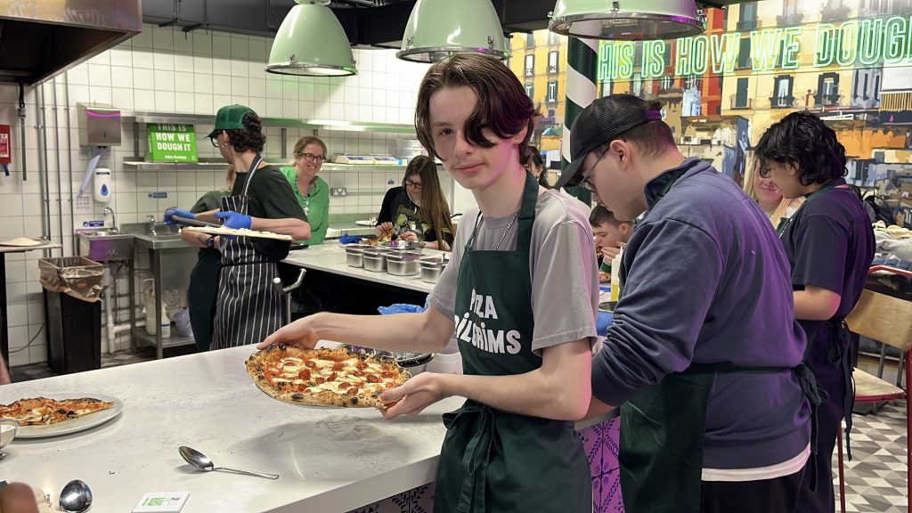 Young man holds up a freshly baked pizza. He is stood at a counter in a colorful training kitchen.