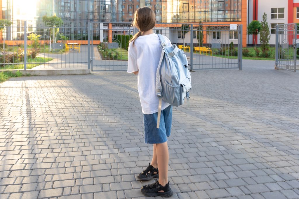 A schoolgirl wearing denim shorts and a white t-shirt with briefcases on her shoulders. 