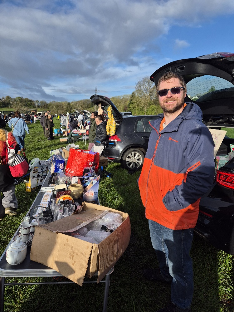 Matt, a man wearing a jacket, is stood in front of his car. In front of him there is a table with items for sale.