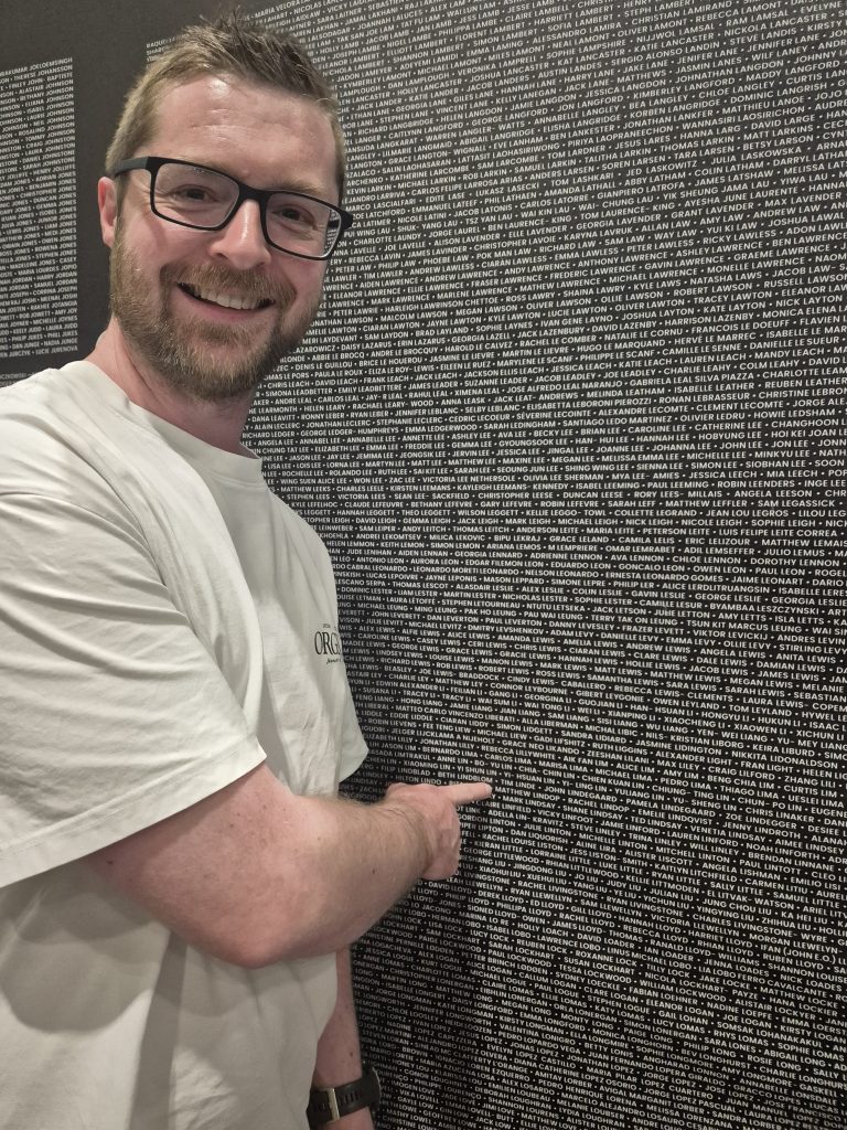 Matt, a man wearing a tshirt, is pointing at his name on the wall of names of runners who completed the london marathon.