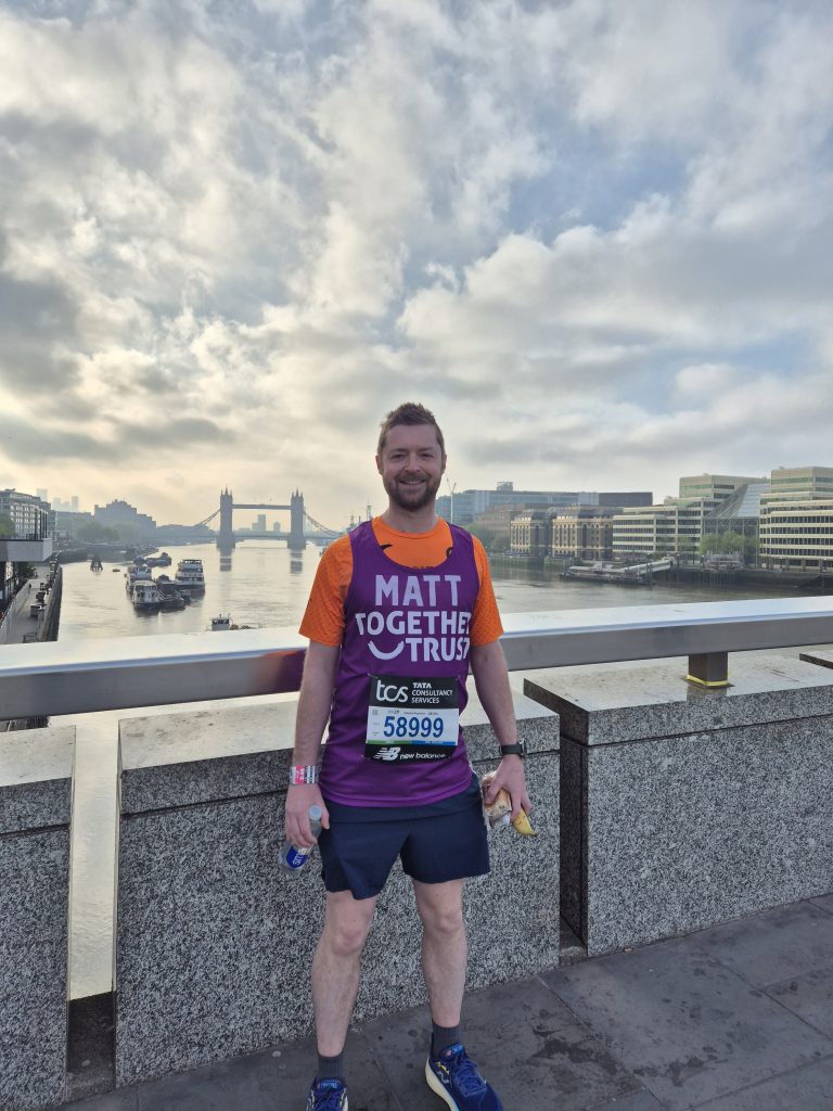 A man is posing in front of the london skyline. He is wearing a together trust running vest. Behind him you can see the silhouette of the Tower Bridge.
