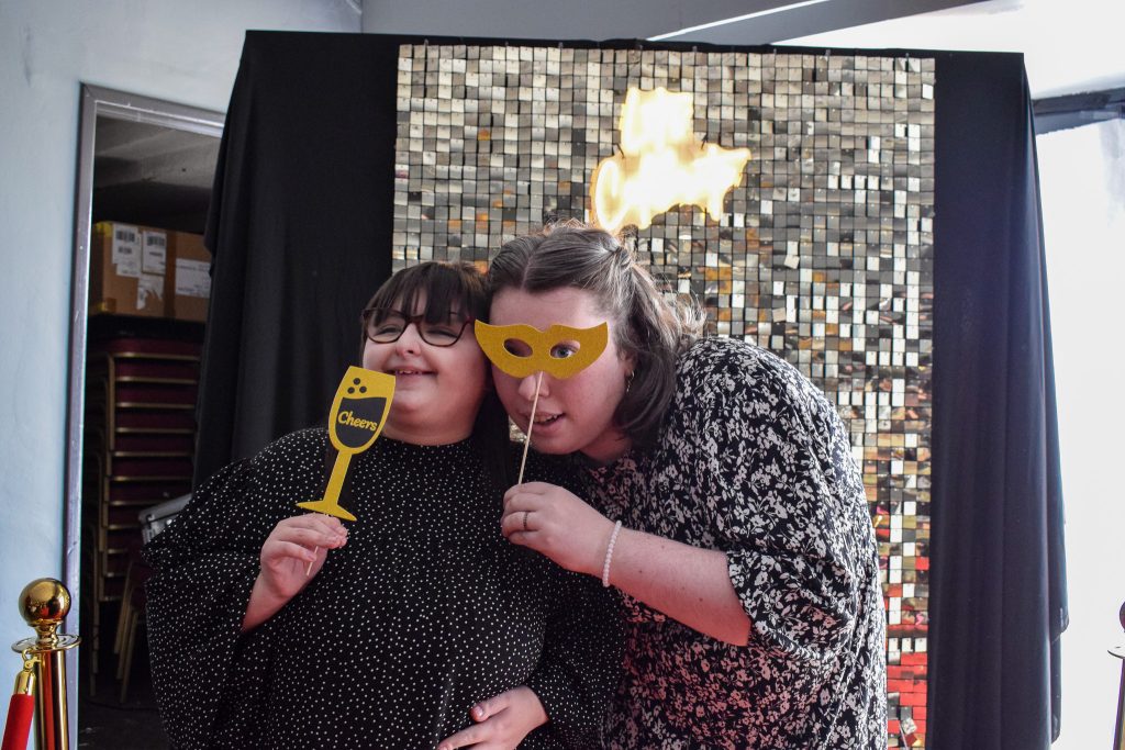 Two women pose in front of a glitter wall by holding up props - a champagne glass and a fancy mask.