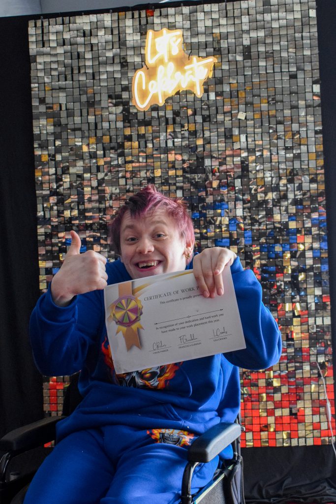 A young man in a wheelchair is giving the camera a thumbs up while holding his diploma. He is posing in front of a glittery wall. 