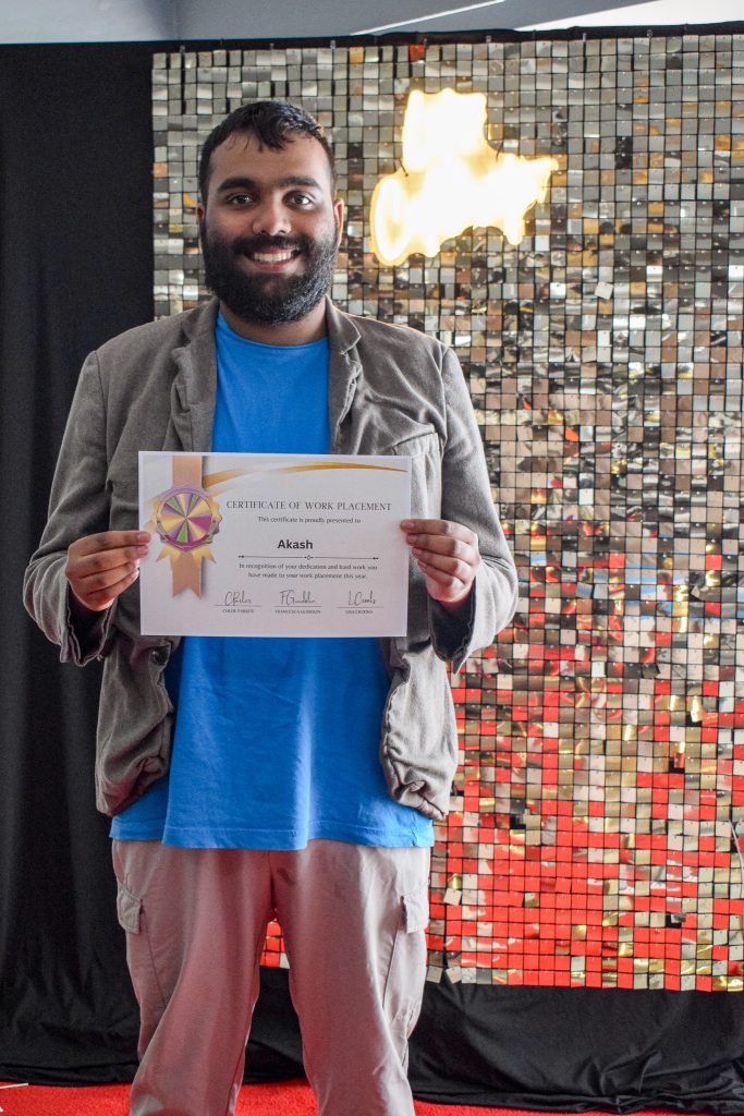 A young man is posing in front of a glittery wall with his diploma. 
