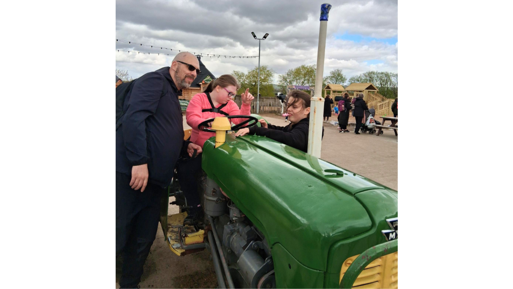 A dad and his two daughters, one of the girls is sat on a green tractor while the other reaches over to touch the steering wheel.