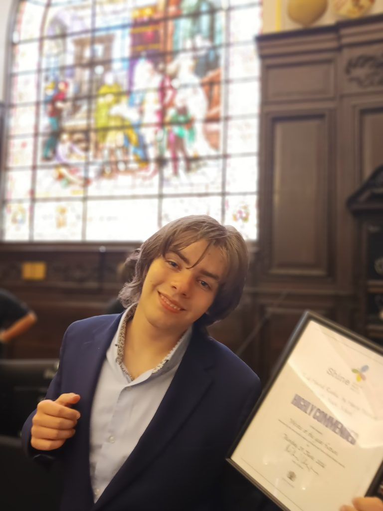 Harry is smiling at the camera holding his award. He is stood in a beautiful room with wood panneling and stained glass windows.