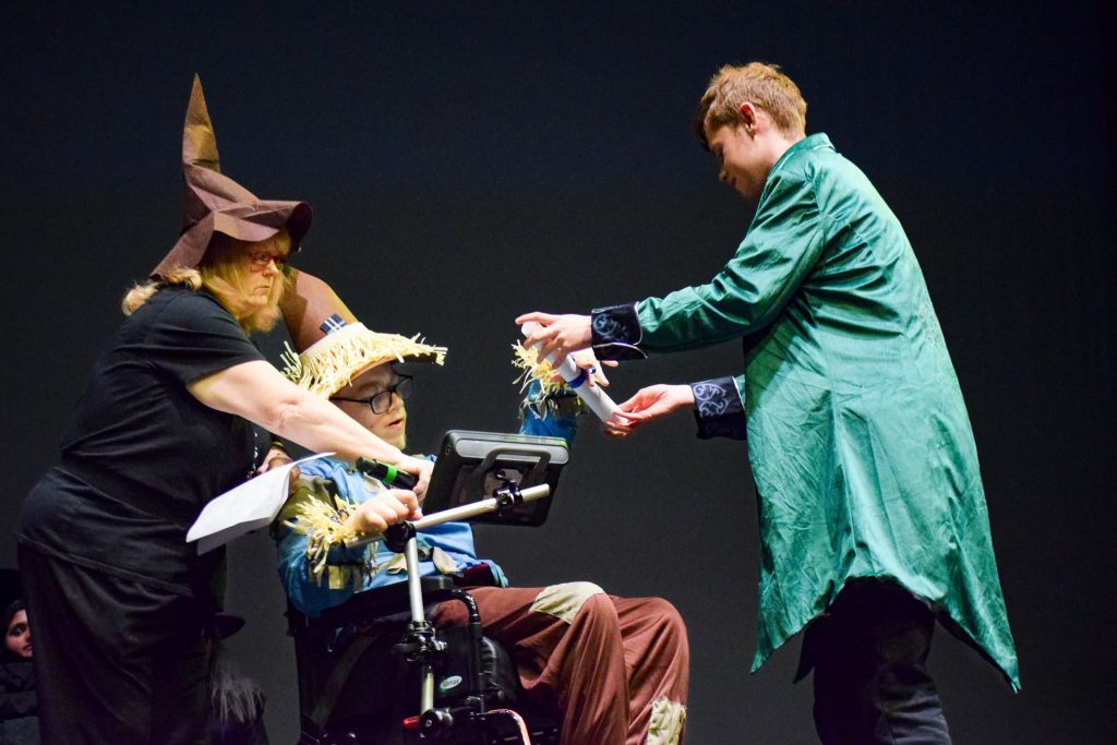 Two students acting in the Wizard of Oz play. One is in a wheelchair, and dressed like the scarecrow. The other student is wearing a  Wizard of Oz costume. 