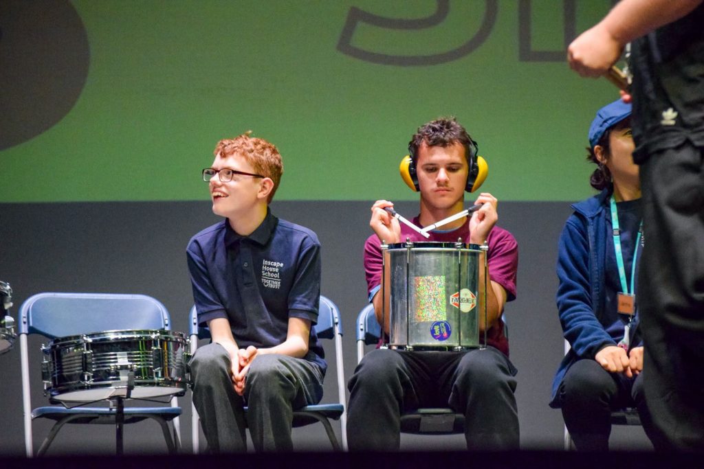 Two boys are sat on plastic chairs on the stage ready to perform on drums. 