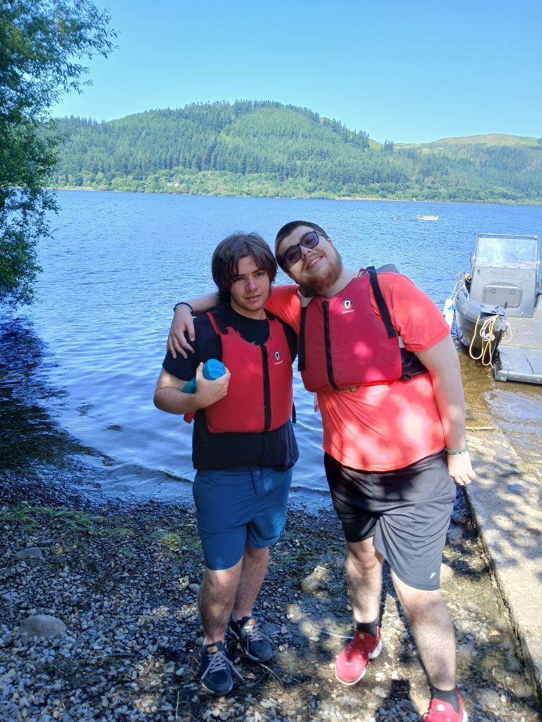 two students wearing life vests pose on the shore in front of the lake