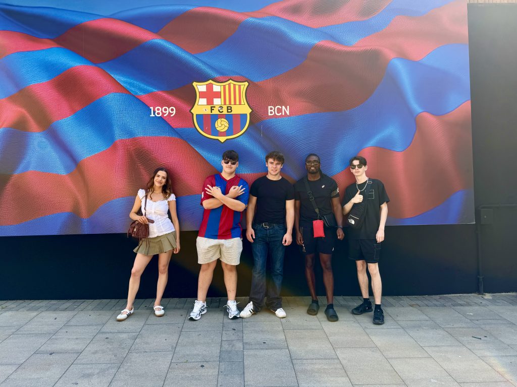A group of students and staff member posing in front of the barcelona football club sign. 