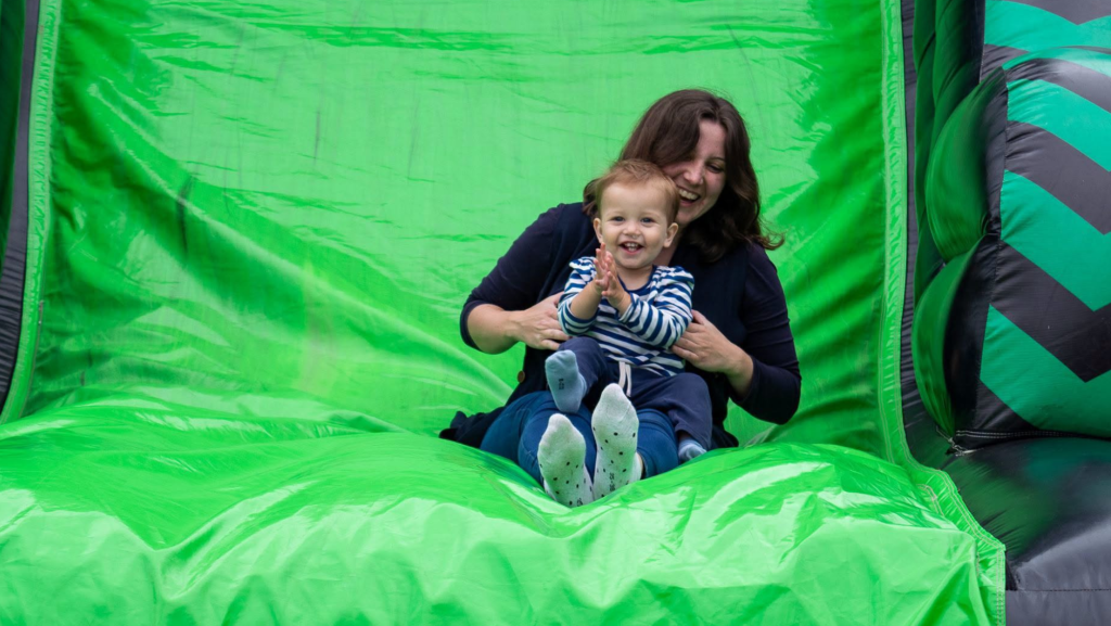 A woman holding a small child sliding down an inflatable slide. The small child is clapping and smiling.