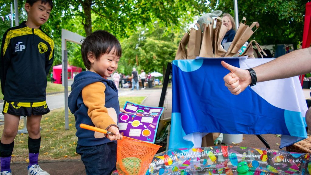 A young child is smiling holding net playing at one of the stalls. The person running the stall is doing the 'thumbs up' pose.