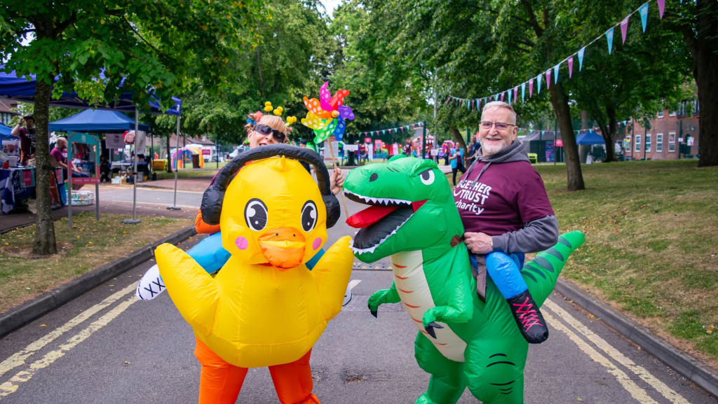 A photo of colleagues wearing blow up fancy dress outfits. The person on the left is dressed as a duck wearing headphones. The person on the right is dressed up as a dinosaur. Both are smiling at the camera.