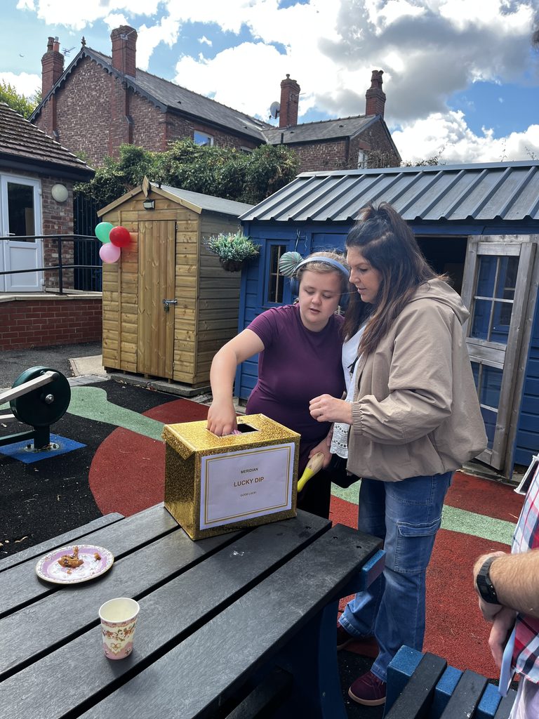 A teen girl, stood next to an woman as they both look at a lucky dip box, about to pick a surprise present out.