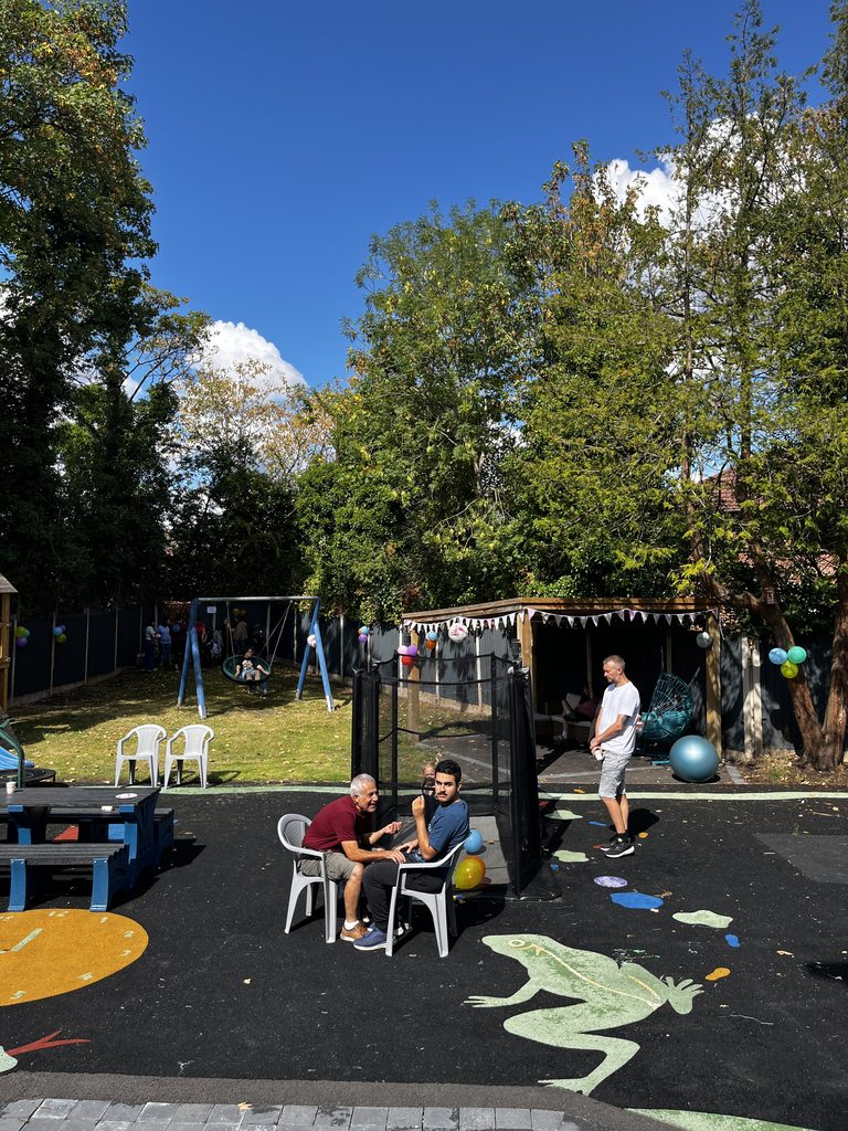 A wide shot of the garden that has a wooden gazeebo, an accessible basket swing, soft tarmac. with frogs painted on. In the garden there are 3 children and two adults enjoying the space.