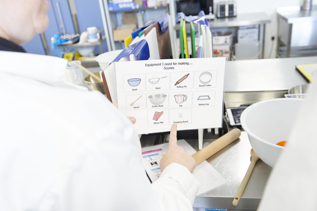 A student is pointing at an instruction sheet for making scones that details what equipment he will need. 