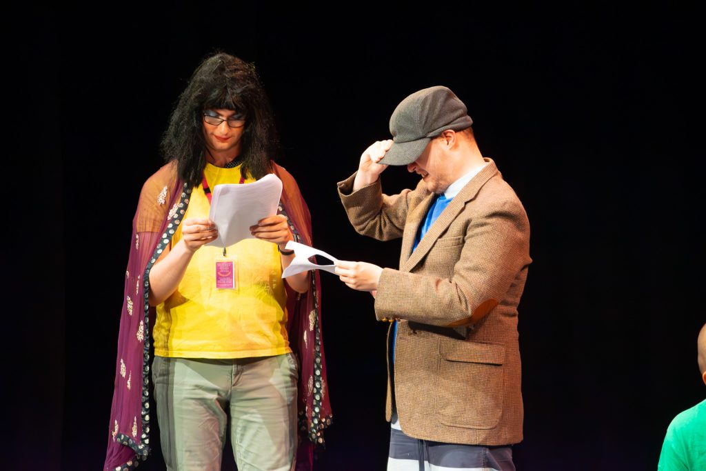two student actors on a stage: one wearing a wig and a cape, while the other is dressed in an old fashioned way.